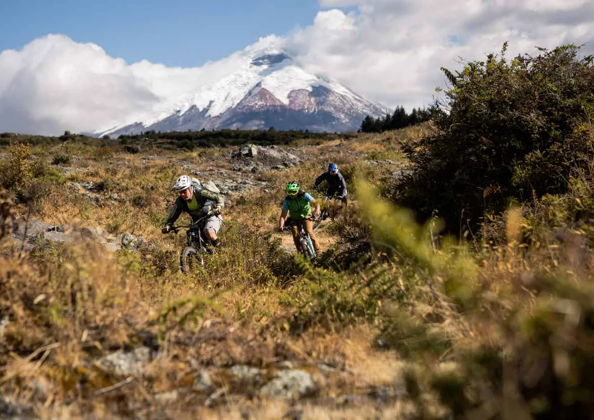 Three mountain bikers descending a rocky trail below the snow-capped Cotopaxi volcano