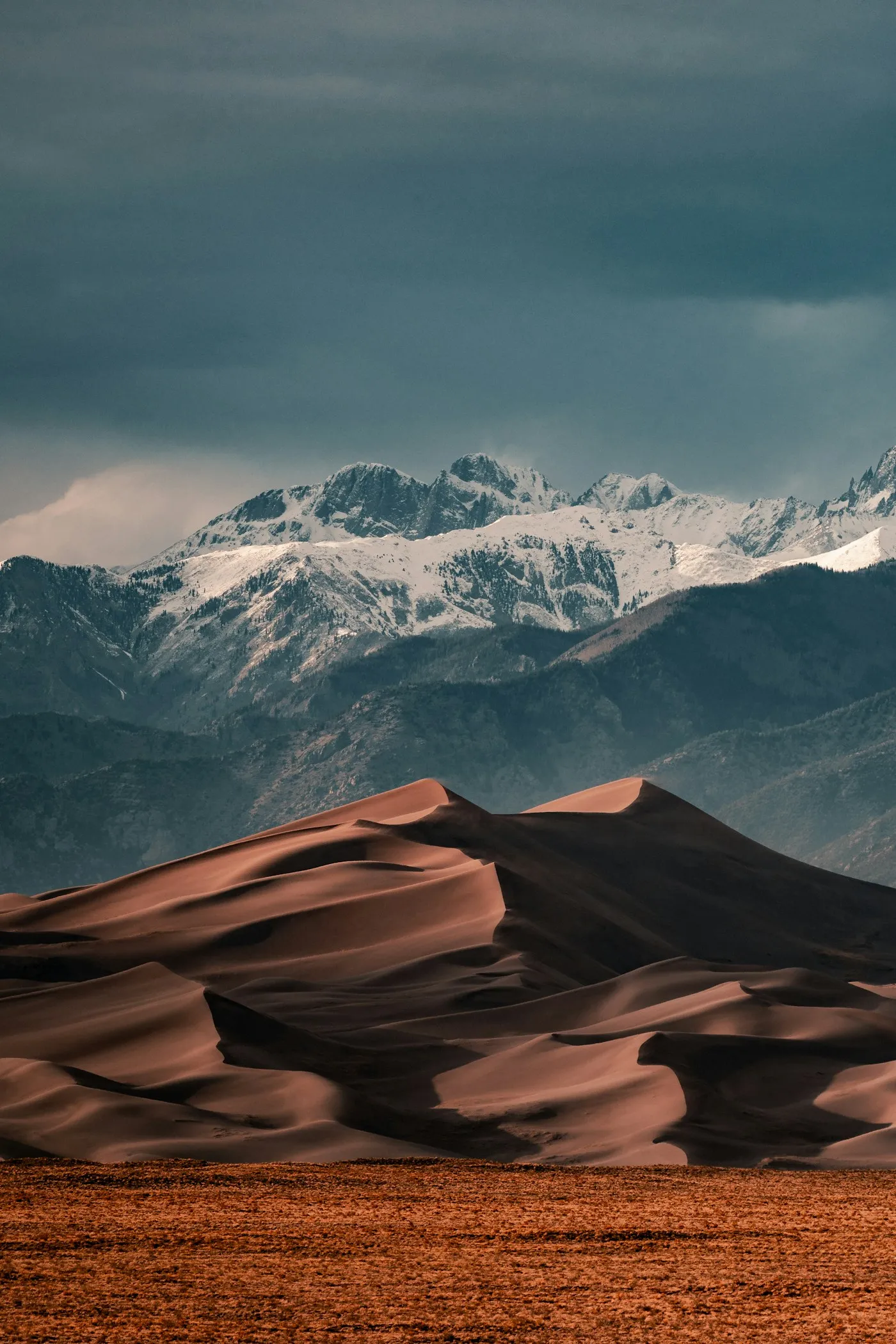 Orange sand dunes in front of snow-capped mountains under a storm sky